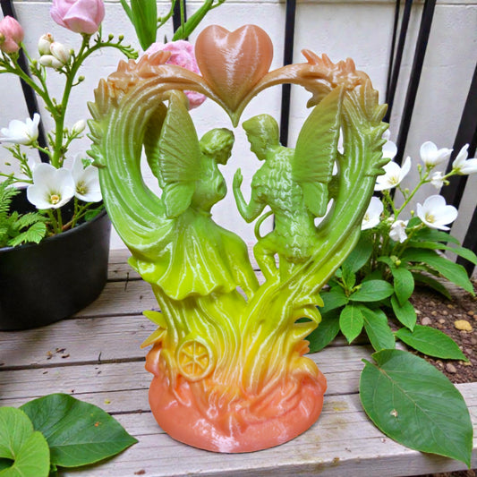 Gradient orange-to-green angel couple heart sculpture displayed outdoors among green leaves and white flowers