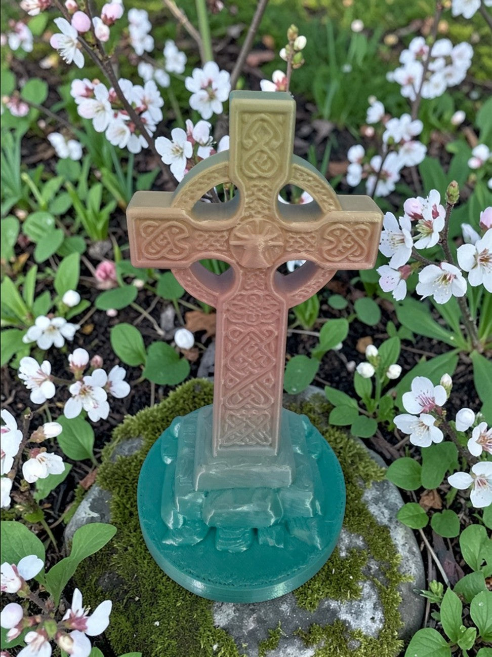3D printed Celtic Cross sculpture with intricate knotwork and Christian motif, standing outdoors on moss with white flowers.