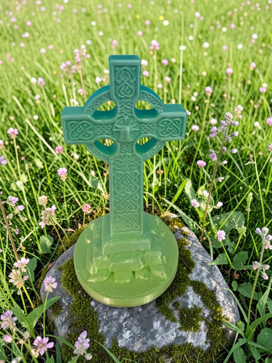 Close-up front view of 3D printed detailed Celtic Cross sculpture in green tones on stone surface with small flowers.